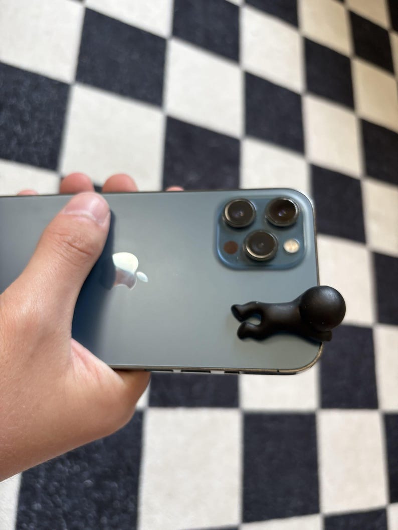Puede incluir: Un smartphone de color verde azulado con una figura negra estilizada adjunta. El tel&eacute;fono tiene una c&aacute;mara de triple lente y el logotipo de Apple. La figura es una peque&ntilde;a forma humana negra abstracta con una cabeza redondeada.