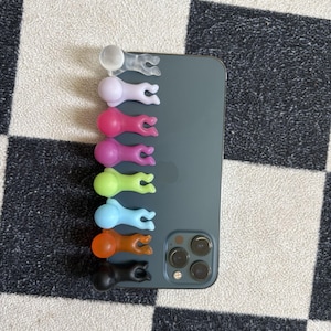 Puede incluir: Una colecci&oacute;n de coloridos agarres para tel&eacute;fono en varios colores, incluyendo transparente, lavanda, rosa, verde, azul, naranja y negro. Los agarres tienen forma de peque&ntilde;as figuras y est&aacute;n adheridos a la parte trasera de un smartphone gris oscuro.