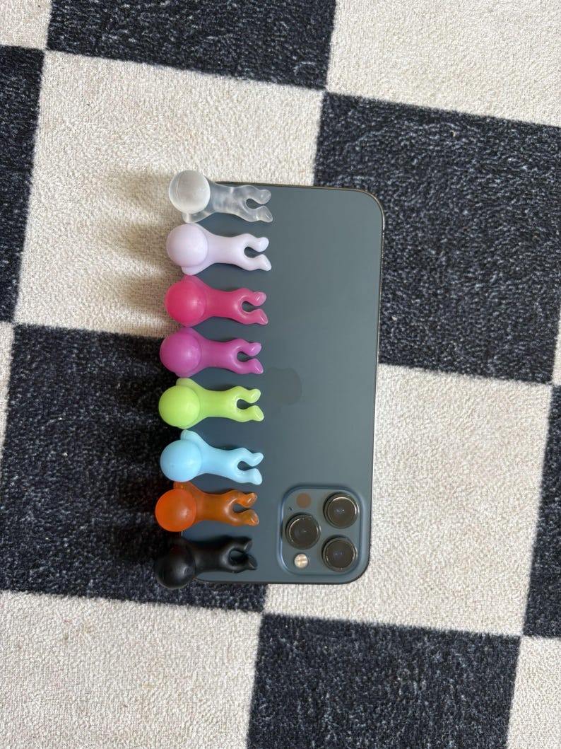 Puede incluir: Una colecci&oacute;n de coloridos agarres para tel&eacute;fono en varios colores, incluyendo transparente, lavanda, rosa, verde, azul, naranja y negro. Los agarres tienen forma de peque&ntilde;as figuras y est&aacute;n adheridos a la parte trasera de un smartphone gris oscuro.