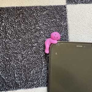 May include: A pink figurine in the shape of a person is perched on the edge of a black smartphone. The figurine is made of a soft material and has a simple, rounded design. The background is a black and white checkered pattern.