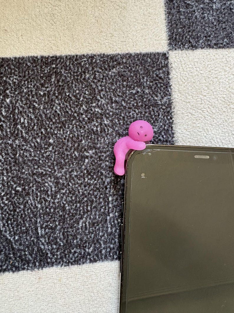 May include: A pink figurine in the shape of a person is perched on the edge of a black smartphone. The figurine is made of a soft material and has a simple, rounded design. The background is a black and white checkered pattern.