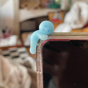 May include: A light blue figurine in the shape of a person is perched on the edge of a smartphone. The figurine has a rounded head and body with small facial features. The phone is in a clear case.