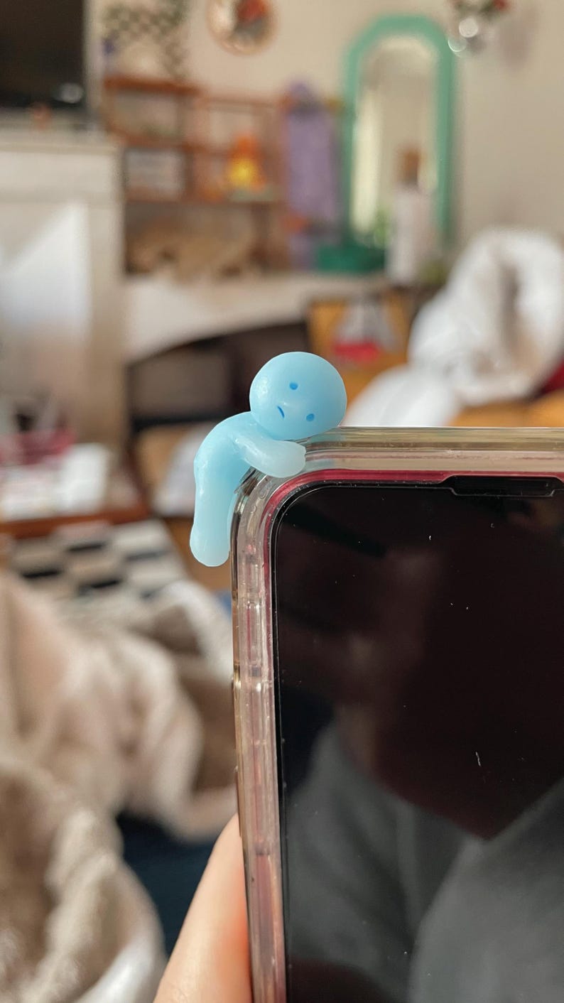 May include: A light blue figurine in the shape of a person is perched on the edge of a smartphone. The figurine has a rounded head and body with small facial features. The phone is in a clear case.
