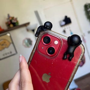 May include: Red smartphone with a clear case, featuring two small black figures clinging to the top edge. The phone has a triple-lens camera and a gold Apple logo. The case is transparent, showing the phone's red back.