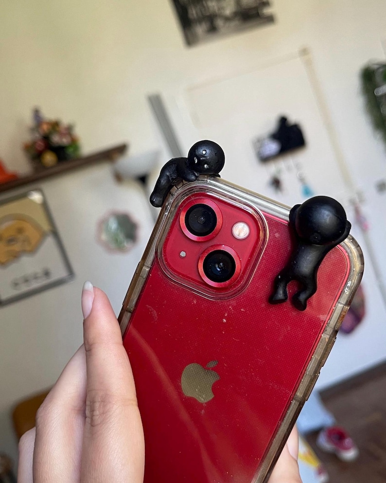 May include: Red smartphone with a clear case, featuring two small black figures clinging to the top edge. The phone has a triple-lens camera and a gold Apple logo. The case is transparent, showing the phone's red back.