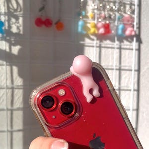 Puede incluir: Un tel&eacute;fono inteligente rojo con una funda transparente, con una peque&ntilde;a figura rosa adherida a la parte superior. Se ven las lentes de la c&aacute;mara y el logotipo de Apple del tel&eacute;fono. En el fondo, pendientes colgantes de colores cuelgan de una rejilla blanca.