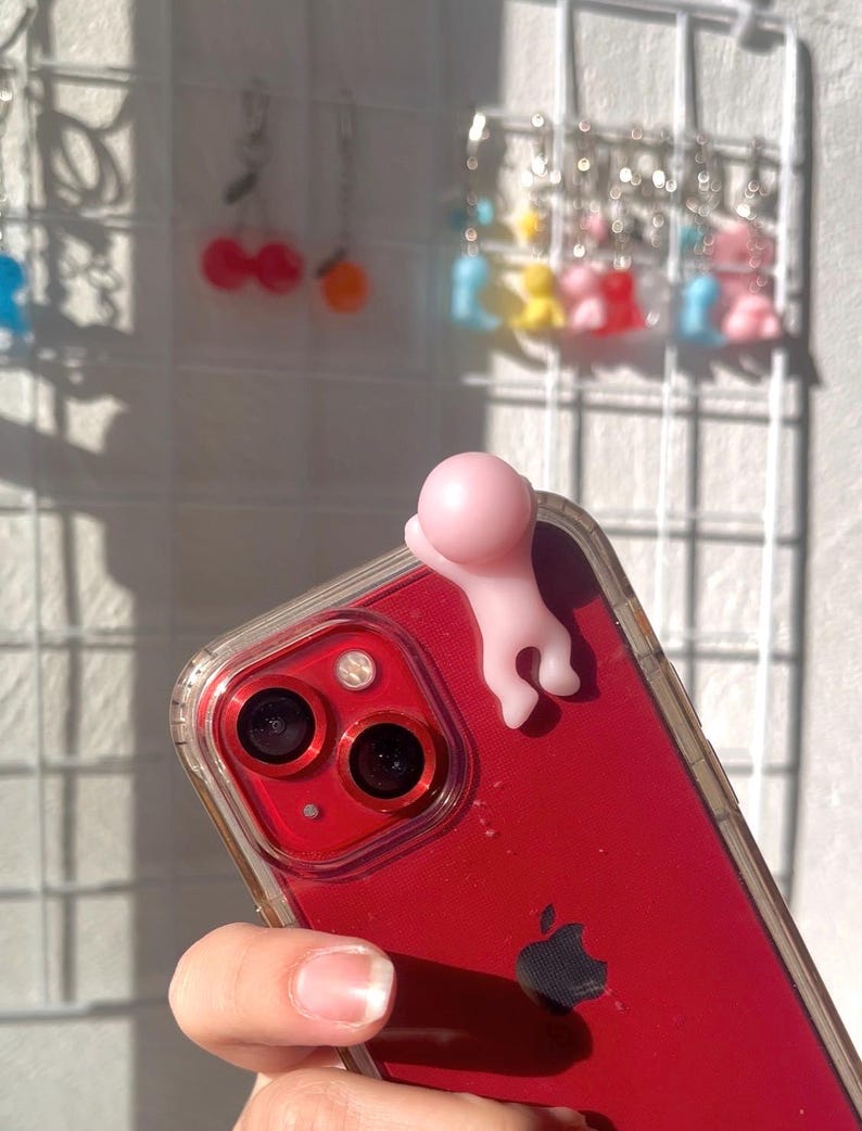 Puede incluir: Un tel&eacute;fono inteligente rojo con una funda transparente, con una peque&ntilde;a figura rosa adherida a la parte superior. Se ven las lentes de la c&aacute;mara y el logotipo de Apple del tel&eacute;fono. En el fondo, pendientes colgantes de colores cuelgan de una rejilla blanca.