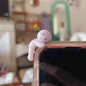 May include: A light purple phone charm in the shape of a small person, perched on the edge of a smartphone. The charm has a simple, minimalist design with a slightly sad expression. The phone is in a clear case.