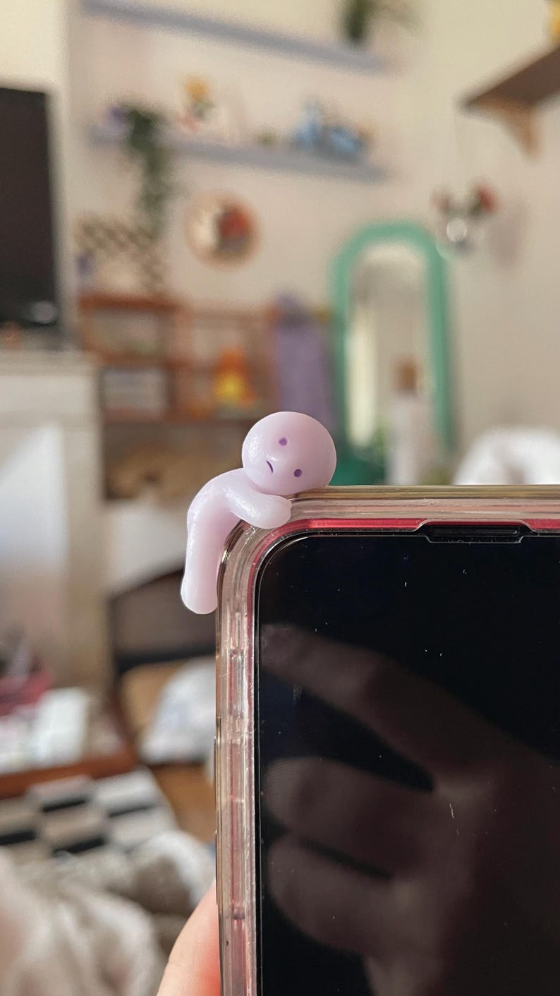 May include: A light purple phone charm in the shape of a small person, perched on the edge of a smartphone. The charm has a simple, minimalist design with a slightly sad expression. The phone is in a clear case.