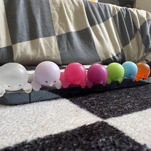 Puede incluir: Una fila de peque&ntilde;as figuras coloridas con forma de foca en varios colores, incluyendo transparente, rosa, magenta, verde, azul, naranja y negro. Las figuras est&aacute;n dispuestas sobre un objeto rectangular gris oscuro, probablemente una funda de tel&eacute;fono. El fondo es un patr&oacute;n de cuadros blanco y negro.
