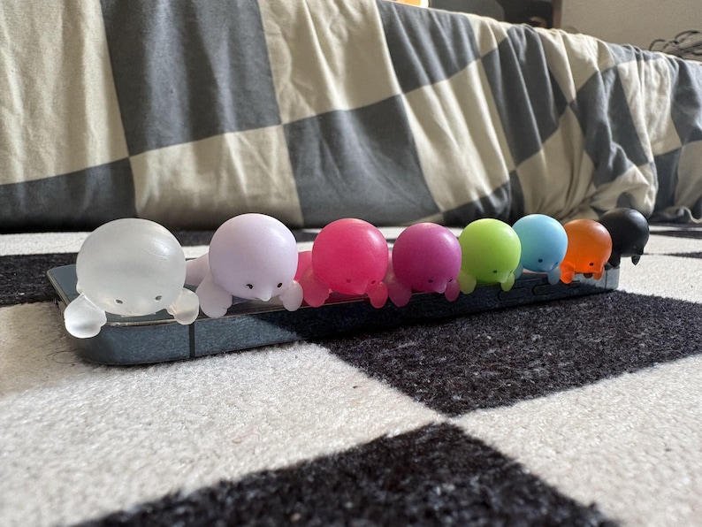 Puede incluir: Una fila de peque&ntilde;as figuras coloridas con forma de foca en varios colores, incluyendo transparente, rosa, magenta, verde, azul, naranja y negro. Las figuras est&aacute;n dispuestas sobre un objeto rectangular gris oscuro, probablemente una funda de tel&eacute;fono. El fondo es un patr&oacute;n de cuadros blanco y negro.