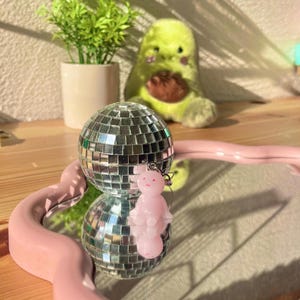 Peut inclure: Une boule disco miroir est posée sur un plateau rose en forme de cœur, reflétant son environnement. Un petit porte-clés figurine rose est suspendu à la boule. En arrière-plan, une peluche avocat et une petite plante en pot sont visibles.