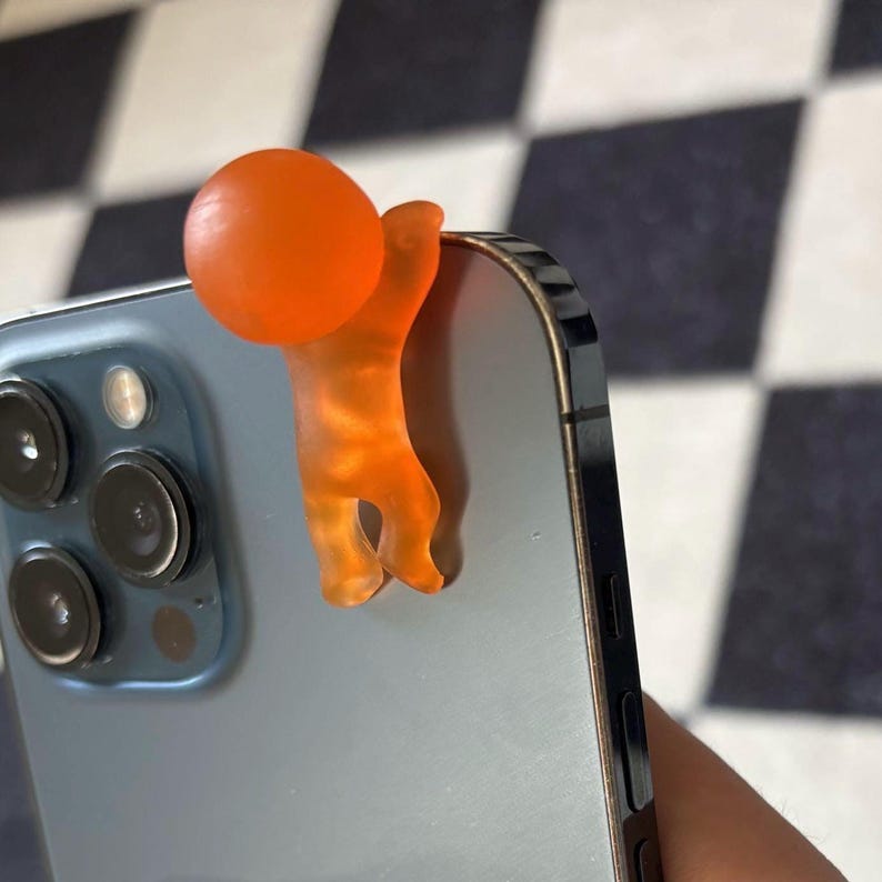 Puede incluir: Un accesorio de tel&eacute;fono naranja con forma de persona agarrada al borde de un tel&eacute;fono. El accesorio tiene una cabeza redonda y un cuerpo con extremidades. El tel&eacute;fono es de color azul claro.