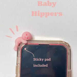 May include: A pink phone accessory shaped like a baby, attached to a phone case. The text "Baby Hippers" is above the phone. The phone screen has the text "Sticky pad included".