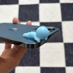 Puede incluir: Un dije de tel&eacute;fono con forma de ballena azul claro apoyado en un tel&eacute;fono inteligente azul. El dije es una peque&ntilde;a ballena estilizada con cabeza y cuerpo redondeados. El tel&eacute;fono tiene un marco plateado y una lente de c&aacute;mara negra.