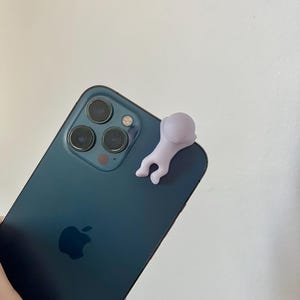 Puede incluir: Un tel&eacute;fono inteligente de color verde azulado con una peque&ntilde;a figura de color p&uacute;rpura claro adjunta. El tel&eacute;fono tiene tres lentes de c&aacute;mara y el logotipo de Apple. La figura tiene la forma de una persona.