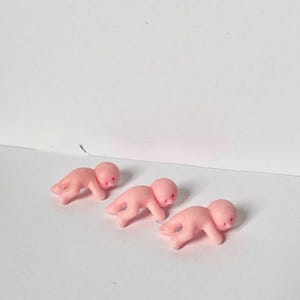 May include: Three small, pink plastic baby figurines in a crawling position. Each has a rounded head and simple facial features. The figurines are arranged on a white surface.