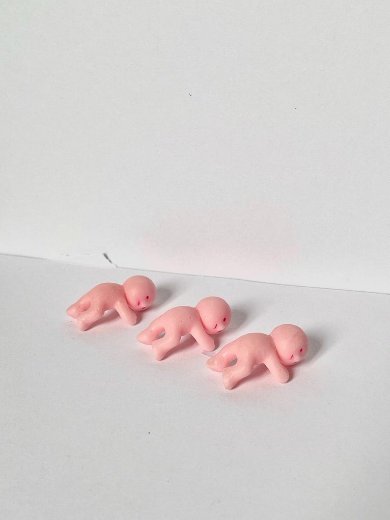 May include: Three small, pink plastic baby figurines in a crawling position. Each has a rounded head and simple facial features. The figurines are arranged on a white surface.