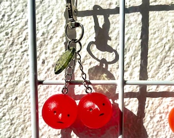 May include: A red cherry keychain with a silver clasp and chain. The keychain features two red cherries with small, sad faces and a green leaf charm. The keychain is hanging on a white grid.