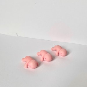 May include: Three small, pink, baby-shaped figurines are arranged on a white surface. Each figurine is lying down, with arms and legs slightly extended. The figures have a smooth, rounded appearance.