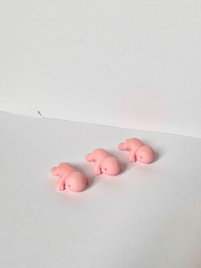 May include: Three small, pink, baby-shaped figurines are arranged on a white surface. Each figurine is lying down, with arms and legs slightly extended. The figures have a smooth, rounded appearance.