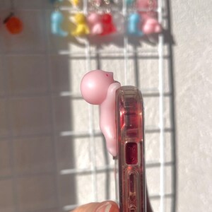 Puede incluir: Un agarre de tel&eacute;fono rosa con forma de peque&ntilde;a figura est&aacute; adherido a una funda transparente con detalles rojos. El agarre est&aacute; en el lateral del tel&eacute;fono. En el fondo, una rejilla blanca muestra varios amuletos coloridos.