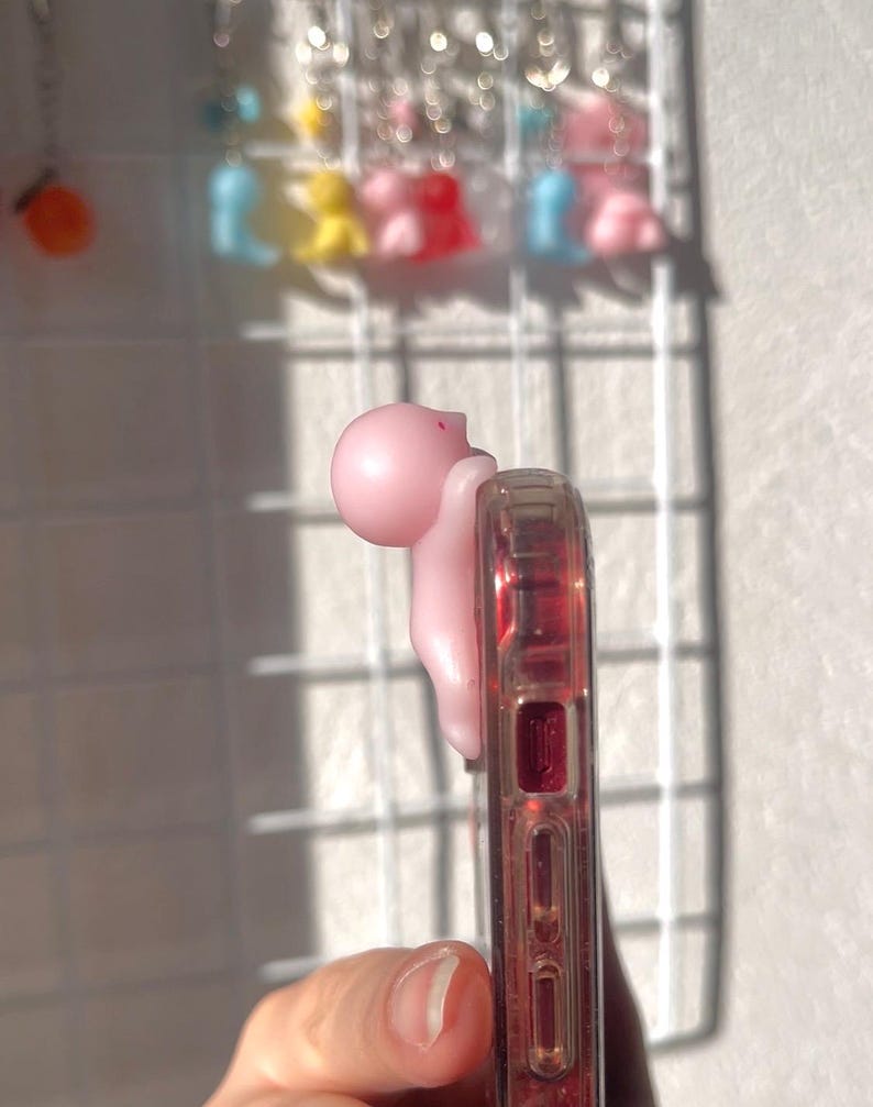 Puede incluir: Un agarre de tel&eacute;fono rosa con forma de peque&ntilde;a figura est&aacute; adherido a una funda transparente con detalles rojos. El agarre est&aacute; en el lateral del tel&eacute;fono. En el fondo, una rejilla blanca muestra varios amuletos coloridos.