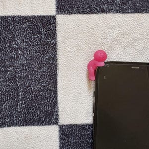 May include: A pink, stylized figure is perched on the edge of a black smartphone. The figure has a rounded head and a curved body, appearing to be holding onto the phone. The background features a black and white checkered pattern.