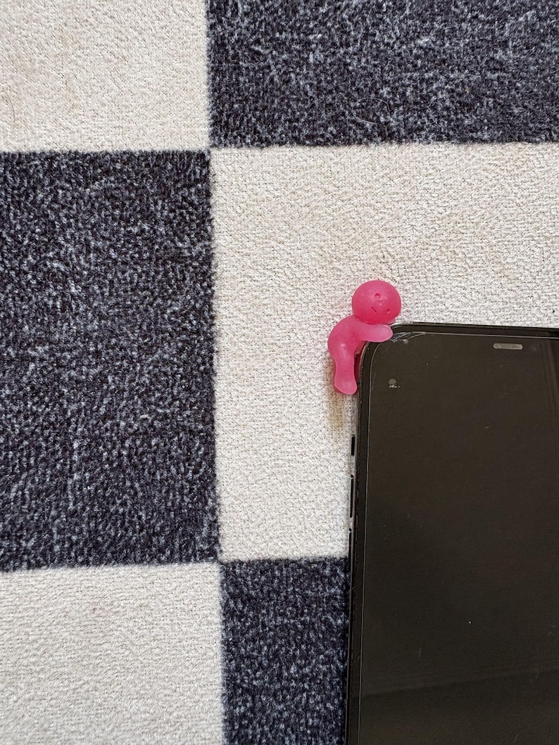 May include: A pink, stylized figure is perched on the edge of a black smartphone. The figure has a rounded head and a curved body, appearing to be holding onto the phone. The background features a black and white checkered pattern.