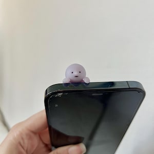 Puede incluir: Una peque&ntilde;a figura de color p&uacute;rpura claro con una cara simple est&aacute; posada encima de un tel&eacute;fono inteligente negro. La figura tiene una cabeza redondeada y brazos peque&ntilde;os. El tel&eacute;fono se sostiene en una mano sobre un fondo blanco liso.