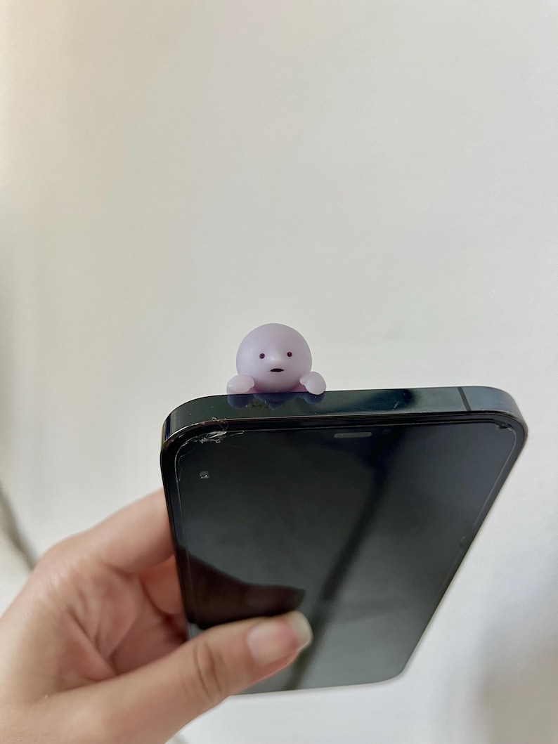 Puede incluir: Una peque&ntilde;a figura de color p&uacute;rpura claro con una cara simple est&aacute; posada encima de un tel&eacute;fono inteligente negro. La figura tiene una cabeza redondeada y brazos peque&ntilde;os. El tel&eacute;fono se sostiene en una mano sobre un fondo blanco liso.