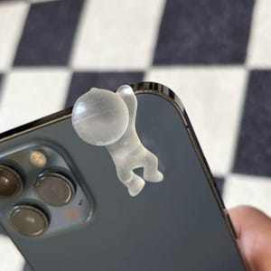 Puede incluir: Un accesorio de tel&eacute;fono de pl&aacute;stico transparente con forma de persona colgando de la esquina de un smartphone gris oscuro. El accesorio tiene una peque&ntilde;a bola y una figura colgante.