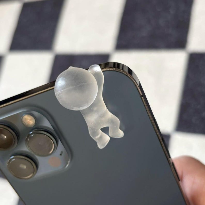 Puede incluir: Un accesorio de tel&eacute;fono de pl&aacute;stico transparente con forma de persona colgando de la esquina de un smartphone gris oscuro. El accesorio tiene una peque&ntilde;a bola y una figura colgante.