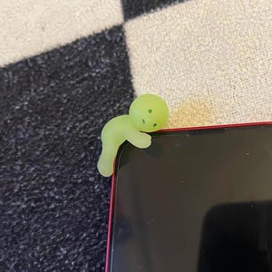 May include: A glowing green phone charm in the shape of a small person is perched on the corner of a black smartphone with a red frame. The charm has a rounded head and a curved body, with small dots for facial features.