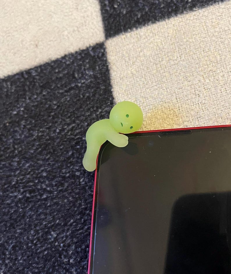 May include: A glowing green phone charm in the shape of a small person is perched on the corner of a black smartphone with a red frame. The charm has a rounded head and a curved body, with small dots for facial features.