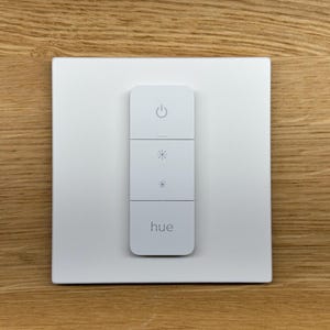 May include: A white wall-mounted smart light switch with a white button and a white 'hue' logo. The switch has four buttons: on/off, brightness up, brightness down, and a button with a sun icon.