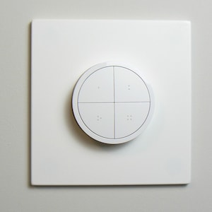 May include: A white, square light switch plate with a circular control. The circular control is divided into four sections by thin black lines, each section containing two small dots. The design is minimalist and modern.