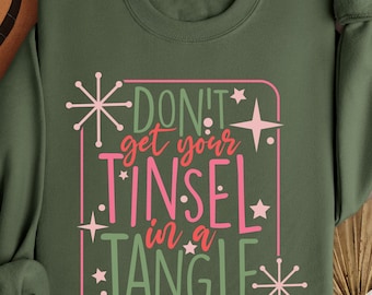 May include: Military green sweatshirt with the text "DON'T get your TINSEL in a TANGLE" in pink and green. The design includes star and snowflake accents. A brown hat is visible in the upper left corner.