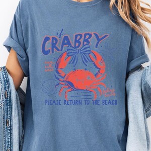 A woman wearing a blue t-shirt with a graphic design of a crab and the text "Please return to the beach" printed on it.