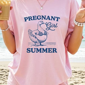 A woman wearing a pink t-shirt with a graphic of a goose and the text "Pregnant Girl Summer" is standing on a beach.