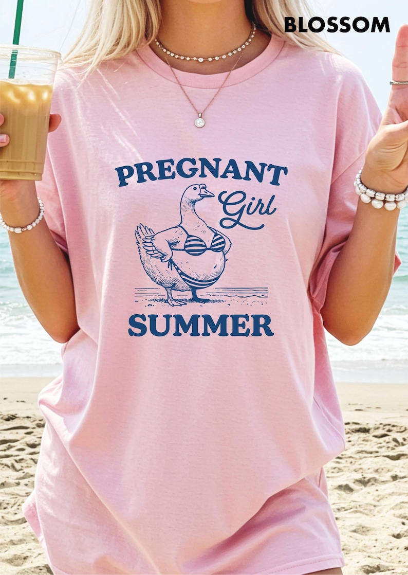 A woman wearing a pink t-shirt with a graphic of a goose and the text "Pregnant Girl Summer" is standing on a beach.