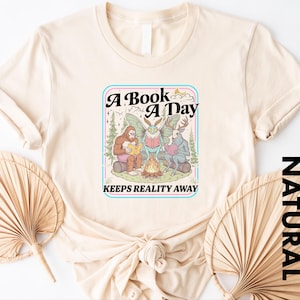 May include: A cream-colored t-shirt featuring a graphic print of a sasquatch, a moth-like creature, and a deer sitting around a campfire reading books. The text reads A BOOK A DAY KEEPS REALITY AWAY. The design includes trees and a crescent moon.