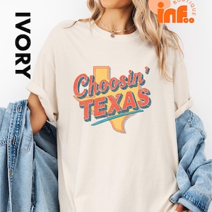 May include: Ivory-colored t-shirt with the text "Choosin' TEXAS" in a retro design. The graphic features a yellow outline of the state of Texas with red and blue text. The shirt is a casual style.