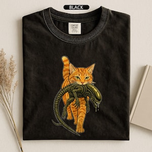 May include: A black crew neck t-shirt featuring a graphic print of an orange tabby cat carrying a green alien creature in its mouth. The shirt has a Comfort Colors brand label at the inner neckline and the text BLACK printed at the top center.