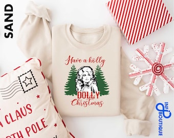 May include: A sand-colored crewneck sweatshirt featuring a graphic print of a person with blonde hair centered between two green pine trees. The text reads Have a holly DOLLY Christmas in red script and block lettering. The word SAND is printed in black.