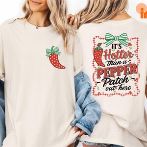 May include: Ivory-colored t-shirt featuring a red chili pepper with white polka dots and a green bow. The back of the shirt displays the text "It's Hotter than a Pepper Patch out here" within a red and white checkered frame.