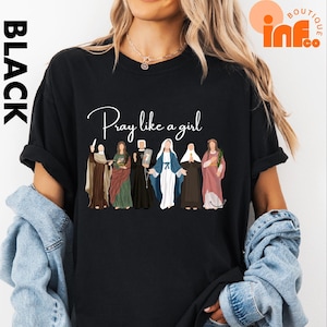 A woman wearing a black t-shirt with a graphic design of various religious figures, including saints and religious icons, along with the text "Pray like a girl".