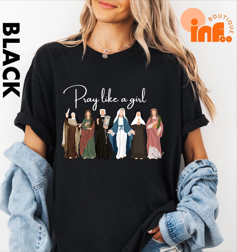 A woman wearing a black t-shirt with a graphic design of various religious figures, including saints and religious icons, along with the text "Pray like a girl".