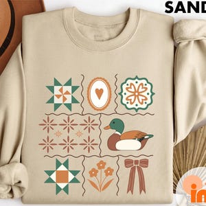 May include: Sand-colored sweatshirt with a grid design featuring a duck, hearts, flowers, and geometric shapes in green, brown, and orange. The word "SAND" is printed at the top. The sweatshirt has a crew neck and long sleeves.