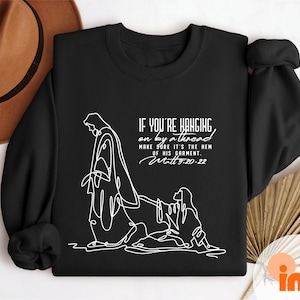 May include: Black crewneck sweatshirt with white line art of a religious figure and text. The text reads "IF YOU'RE HANGING on by a thread MAKE SURE IT'S THE HEM OF HIS GARMENT. Matt 9:20-22". A brown hat is visible in the upper left corner.